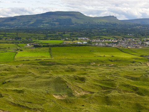 Aerial view of Finner sand dunes, lush green fields, and a distant town nestled beneath a prominent mountain range in County Donegal, Ireland.