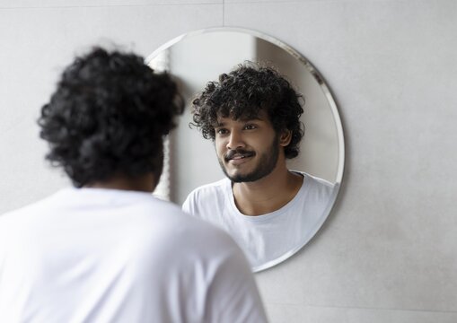 Smiling Young Man With Curly Hair Standing In Front Of Mirror At Home, Engaging In Self Reflection, Enjoying Moment Of Self-acceptance In Cozy Indoor Setting. Home Wellness Concept.