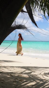 A woman in a flowing beige dress walks along the white sand shore of a tropical beach in Maldives framed by palm fronds.