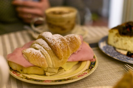 S&aacute;ndwich de croissant relleno de jam&oacute;n y queso derretido, espolvoreado con az&uacute;car impalpable, servido en plato floral con caf&eacute; de fondo.