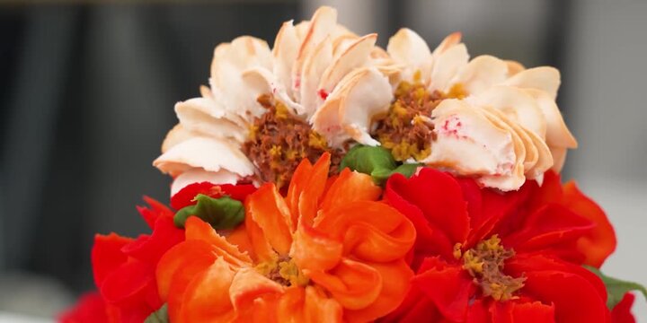 A detailed, macro shot showcasing beautifully piped orange and red buttercream dahlia and zinnia flowers on top of a frosted white cake. The vibrant icing details, delicate textured petals, and piped 