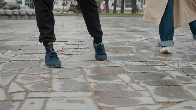 Close up of feet in motion on cobblestone pavement showing person wearing dark pants and blue sneakers with patterned socks walking alongside another person. scene includes visible