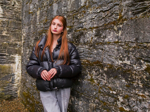 Young woman leans against rugged stone fence, arms together. She wears black puffer jacket and grey pants. Her eyes closed, head tilted back, smiling peacefully. Feels calm and connected with nature