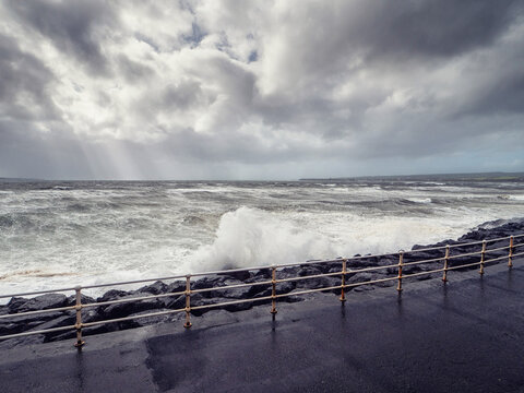 Stormy sea crashes against dark rocks. Waves surge with white foam. Metal railing lines wet foreground. Sky filled with gray clouds. Dramatic nature scene for travel or weather content