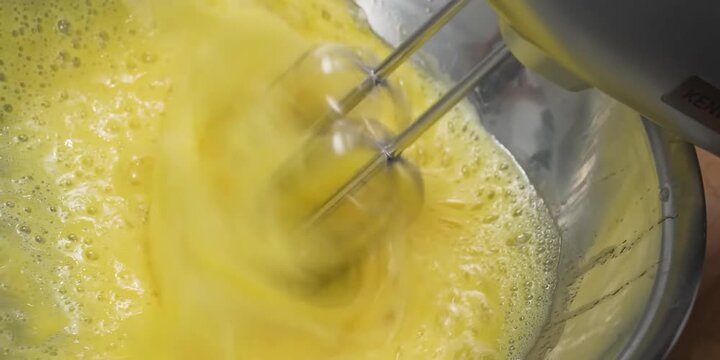 Close-up shot of an electric hand mixer whipping and beating raw yellow eggs in a stainless steel mixing bowl. The motion blur captures the high-speed rotation of the beaters, creating a frothy, bubbl