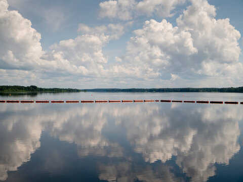 Serene lake mirrors fluffy white clouds overhead. Red buoys line horizon, calm water reflects sky. Peaceful scene ideal for travel, nature, or relaxation themes. Perfect for websites, magazines