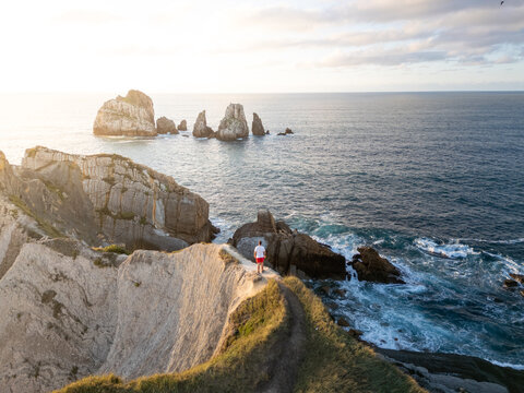 Aerial view of a man standing on a cliff edge overlooking the rugged coastline and dramatic rock formations in the sea at sunset in Liencres, Cantabria, Spain.