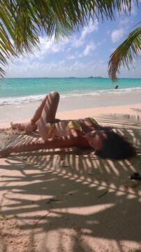 A woman in a yellow bikini relaxes on the sandy shore under palm fronds with ocean waves and clear skies visible on an island in Maldives.