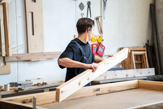 Woodworker preparing materials for antique wooden window frame restoration in woodworking studio, master holding long wooden board, disassembling frame and placing details at workbench in carpentry.