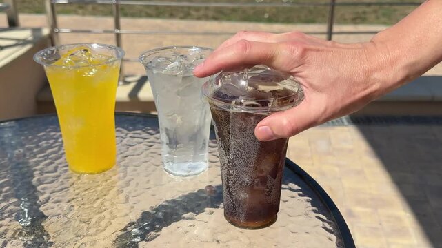 Protective lids are pressed firmly onto three cups of cola, water and a yellow drink, keeping the beverages secure and preventing leaks during transport or outdoor service. Close up.