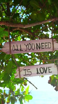 Handcrafted wooden sign with all you need is love text hanging from tree branch with ropes on tropical Maldives beach under bright sunlight.