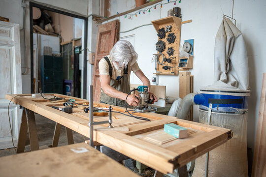 Professional master restorer working in woodworking workshop with electric wood router at workstation, focused crafter using equipment and processing wooden door surface with handheld plunge router.