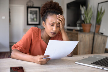 Young African woman holding sheet of paper, looking emotionally exhausted or disappointed due to...