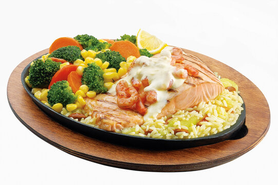 Grilled salmon fillet topped with shrimp and white sauce, served with rice and steamed broccoli, corn, and carrots on a sizzler plate isolated on white.