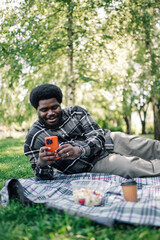 Fototapeta premium Young black man relaxing on picnic blanket in park, using smartphone, enjoying outdoor leisure and technology connection