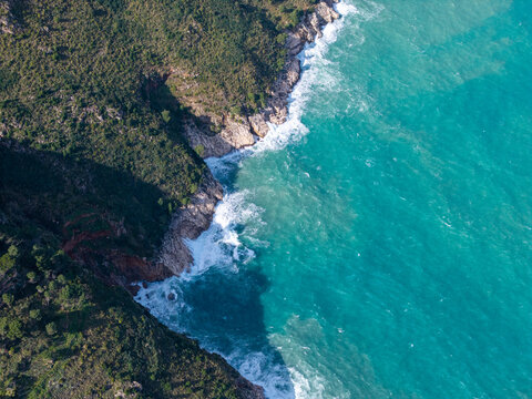 Aerial view of the rugged coastline with turquoise sea waves crashing against rocky cliffs and green Mediterranean vegetation in Castellammare del Golfo, Sicily, Italy.