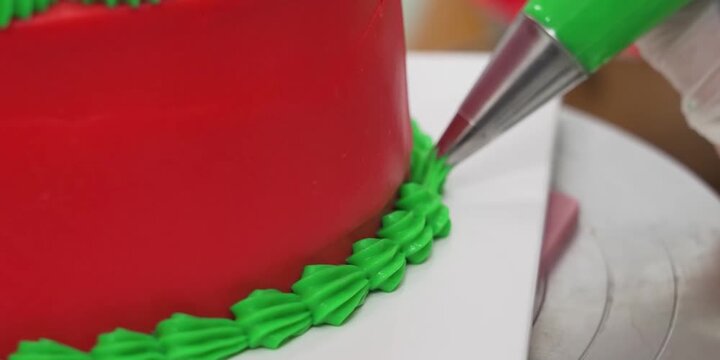 A close-up shot capturing a pastry chef wearing a white glove precisely piping a bright green buttercream shell border onto the bottom edge of a smooth red fondant cake using a star-tipped piping bag.