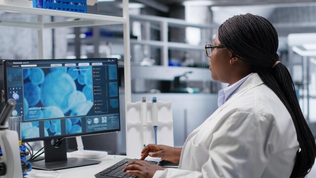 African American woman researcher examines a compound in flasks and containers during laboratory testing, studying molecular content with biotechnology equipment for genetics research.