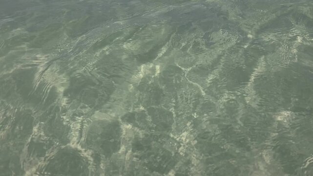 Close-up of crystal-clear shallow water in Montecristi, Dominican Republic, revealing a rocky seabed beneath sunlit caustic patterns. The natural texture and gentle ripples create a calming, abstract 