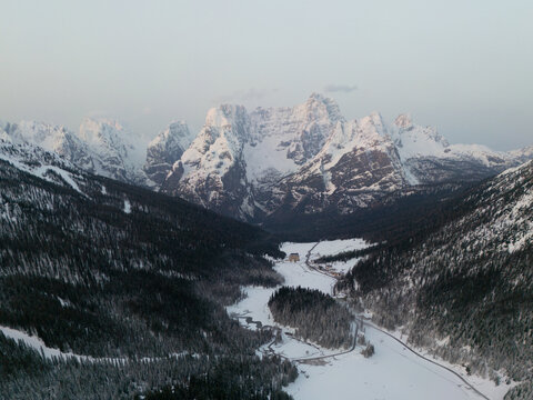 Aerial view of the snow-covered peaks of Dolomiti Bellunesi National Park, with dense pine forests and a frozen valley floor in the winter landscape of Veneto, Italy.