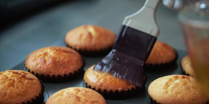 A high-definition, slow-motion, close-up shot capturing a pastry chef using a silicone basting brush to coat freshly baked, warm vanilla cupcakes with a glossy simple syrup glaze. The camera captures 