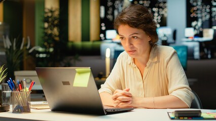 Fototapeta premium Woman at a desk smiles into the webcam during a video call with attentive focus, engaging in a virtual conversation and exchanging ideas with remote participants while working in the office.