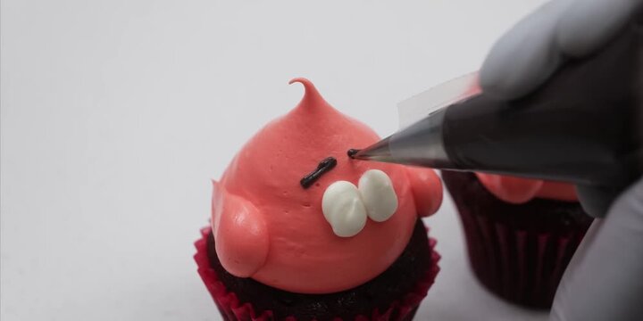close-up shot of a baker using a piping bag with a fine round tip to draw facial expressions on a Patrick Star themed cupcake. The cupcake features bright pink frosting shaped like the cartoon charact