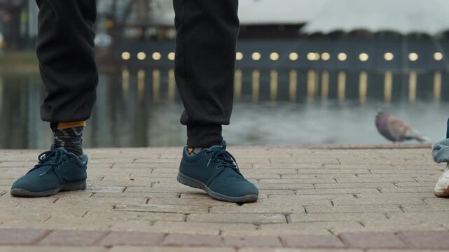 Close up of person feet and legs performing rhythmic footwork on paved ground near water. subject wears dark pants blue sneakers with laces patterned socks visible above ankle while another