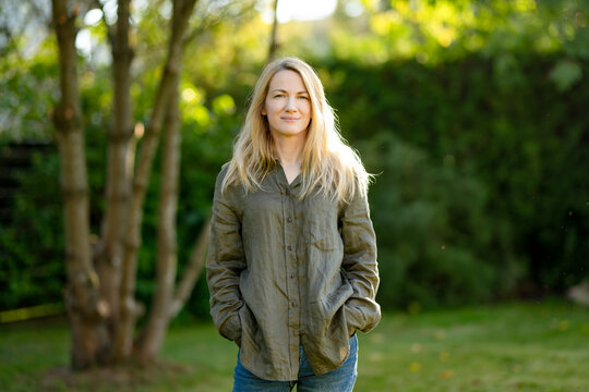 Portrait of a smiling blonde woman outdoors in soft evening sunlight. Relaxed adult woman standing in a green garden with natural backlight and blurred background.