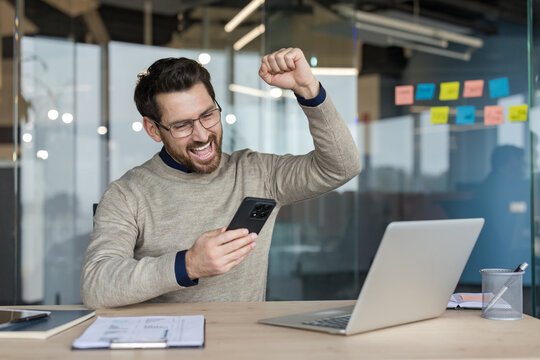 Happy man celebrating great news, triumphantly raising his fist while looking at his smartphone, sitting at an office desk with a laptop and documents