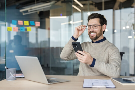 Smiling businessman wearing glasses and holding a mobile phone, sitting at a desk with a laptop, enjoying successful communication and online interaction in a bright corporate workspace