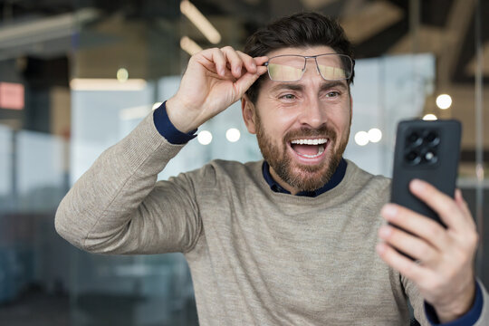 Excited man holding smartphone and lifting his eyeglasses, reacting with joy and triumph to positive news or a successful outcome while working in a modern office environment