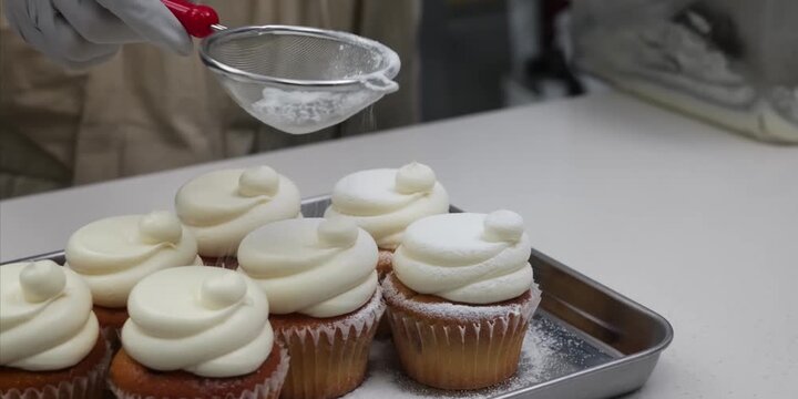 Close-up shot of a pastry chef wearing a protective glove dusting icing sugar through a small metal sieve over vanilla cupcakes topped with smooth, creamy white swirls of buttercream frosting.