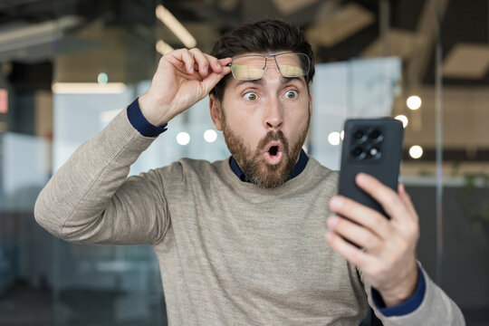 Man with glasses pushed up on his forehead, looking at a smartphone with a wide-eyed, open-mouthed expression of shock and disbelief while receiving unexpected news or content