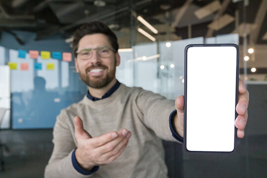 Smiling man in a modern office presenting a mobile phone with a blank white screen, extending his arm and gesturing to the device ideal for app, tech or business mockups