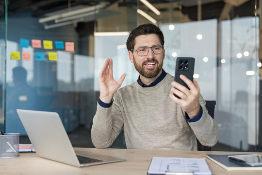 Young businessman communicating remotely via video call on a smartphone, sitting at his modern office desk with a laptop, engaging in a virtual meeting