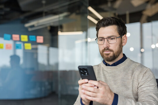 Man in business casual attire and eyeglasses concentrating on his smartphone while working in a modern office environment, connecting with technology and managing tasks
