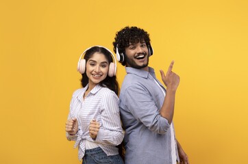Fototapeta premium Smiling Young Asian Woman And Man Enjoying Music While Wearing Headphones, Standing Back To Back In Studio With Bright Yellow Background. Cheerful Emotion Captured In Fun Atmosphere.