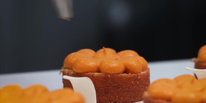 Close-up shot of a pastry chef using a piping bag with a round metal nozzle to decorate a small round sponge cake. The chef carefully squeezes smooth, vibrant orange cream or fruit curd into neat, bul