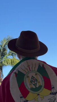 A hat on the head and the flag of Rio Grande do Sul (southern Brazil) across the chest, amidst nature and under a blue sky. Tradition, freedom, and pride in being a *ga&uacute;cho*. Farroupilha Week.