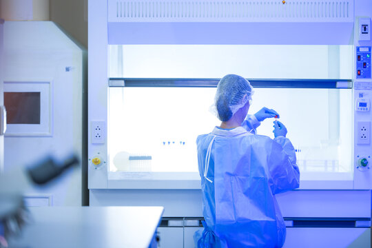 A female microbiologist in a white coat, protective eyewear and a sterile hairnet lifting a gloved hand into the hood opening inside a clean pharmaceutical research laboratory.
