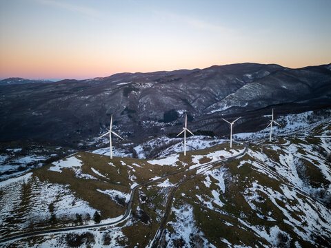 Aerial view of wind turbines at Parco Nazionale dell'Appennino Tosco-Emiliano on a snow-covered mountain ridge during golden hour in Sassalbo, Provincia di Massa e Carrara, Italy.
