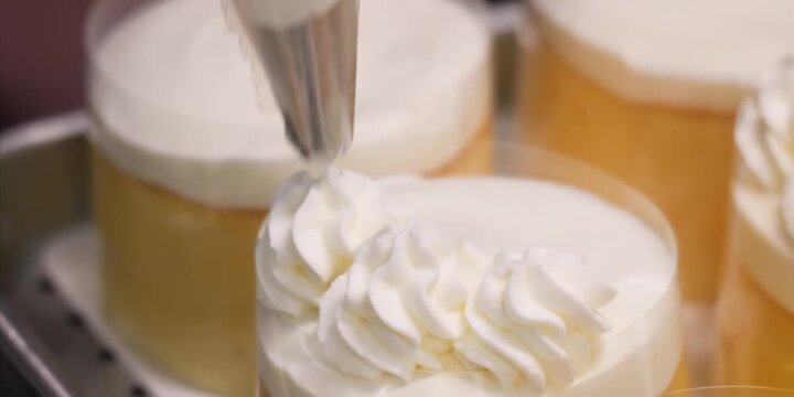 A high-definition, close-up stock shot capturing a professional chef or baker using a piping bag with a star tip nozzle to create perfect, decorative swirls of fluffy white whipped cream on top of ind