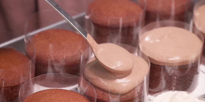 A high-definition, close-up shot capturing a pastry chef using a spoon to neatly spread thick, creamy chocolate mousse over rows of mini sponge cakes wrapped in clear acetate plastic collars. Perfect 