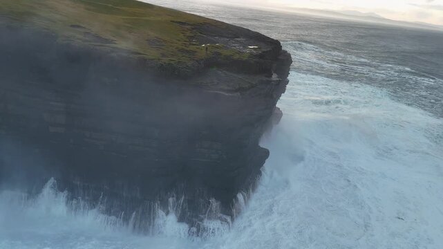 Cinematic drone view of a dark rocky cliff edge and isolated sea stack surrounded by churning white foam along the Wild Atlantic Way