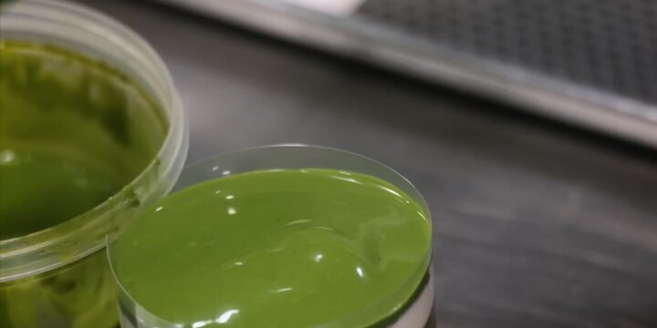 A high-angle, close-up shot capturing thick, glossy matcha green tea sauce being ladled onto a creamy dessert or beverage topping in a professional caf&eacute; kitchen environment. The rich, vibrant green gl
