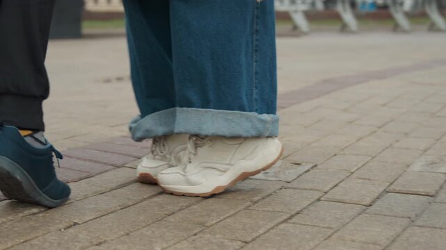 Close up of two people walking showing their feet and lower legs on paved path. One person wears blue jeans with rolled cuffs and white sneakers other has dark pants paired with patterned