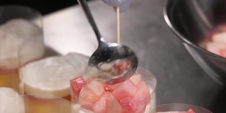 close-up shot of a pastry chef using a metal spoon to carefully arrange fresh, juicy diced peaches on top of a layered cream dessert. The dessert is assembled inside clear plastic collar molds on a pr