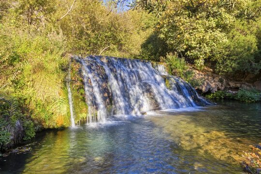 A small waterfall flows over green plants into a clear river under a sunny sky, El Kelaa torrent in Morocco