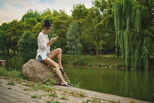 Woman sits on large rock beside calm river focused on smartphone. Green trees surround area time appears to be late afternoon. Relaxed setting in city park